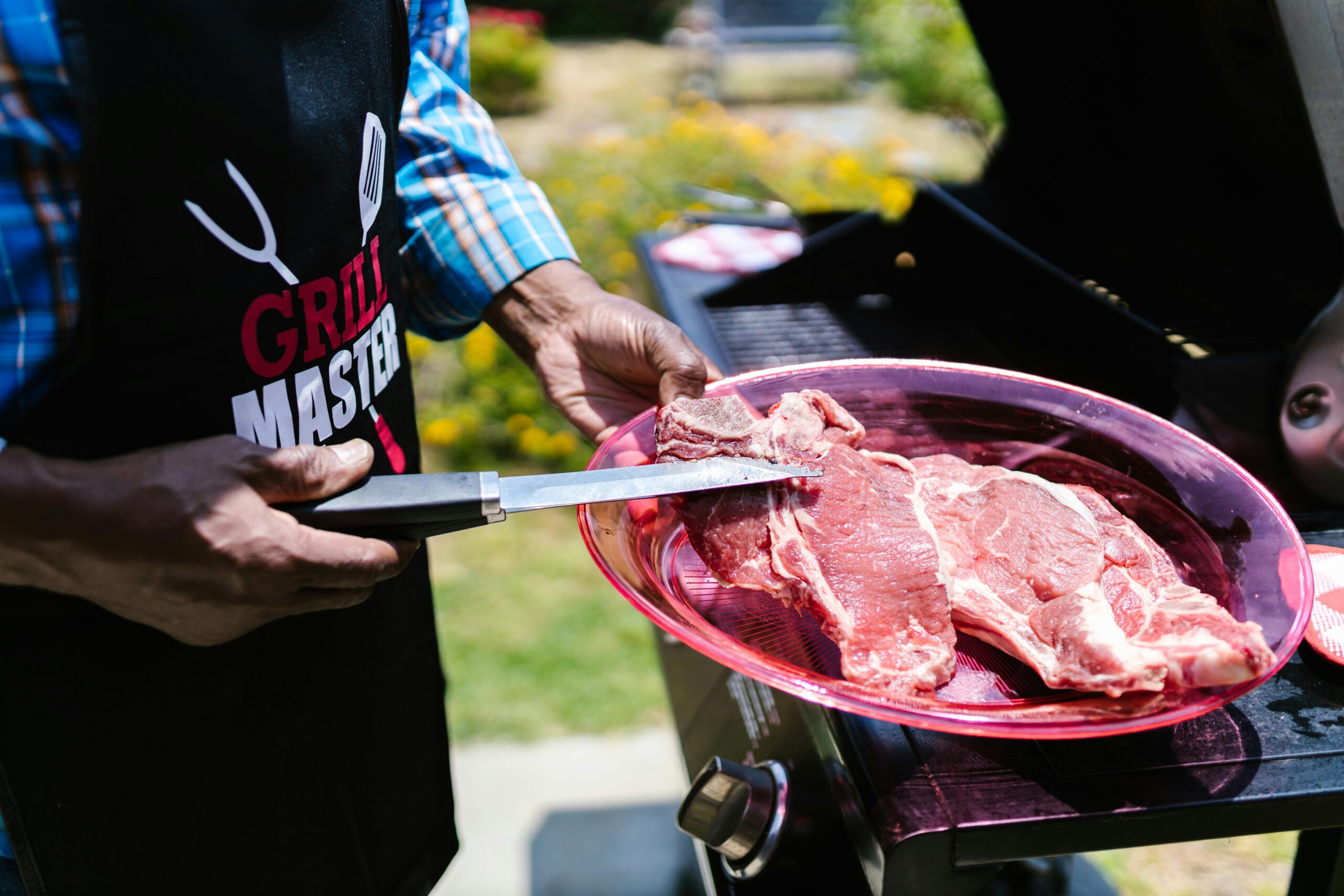 Close-up of a person in a grill master apron preparing raw steaks outdoors on a barbecue grill.
